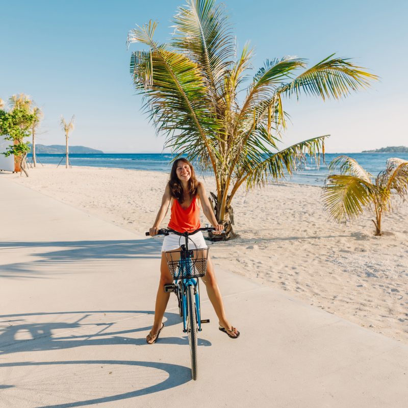 Happy,young,woman,on,blue,bicycle,near,ocean,in,tropical